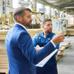 man in a suit with a man with clipboard in a lumber warehouse with a worker talking
