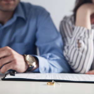 close up of man and woman sitting side by side with paperwork and a pen, and their rings on the table indicating divorce