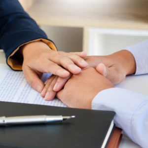 woman in a suit jacket, arm reaching touching a man's hand in a comforting way over paperwork
