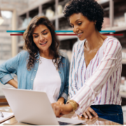 two women in their 30s in their business smiling and discussing business looking at a laptop