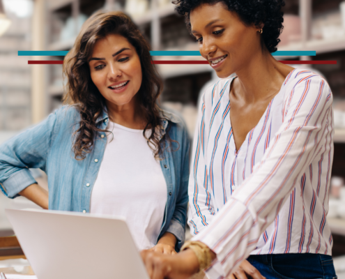 two women in their 30s in their business smiling and discussing business looking at a laptop