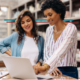 two women in their 30s in their business smiling and discussing business looking at a laptop
