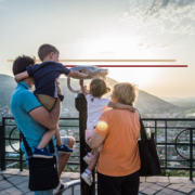 A family of 4 including a father and 2 preschool aged children with their grandmother looking through binoculars over a city in the distance at sunset