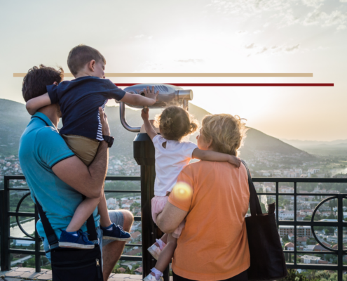 A family of 4 including a father and 2 preschool aged children with their grandmother looking through binoculars over a city in the distance at sunset