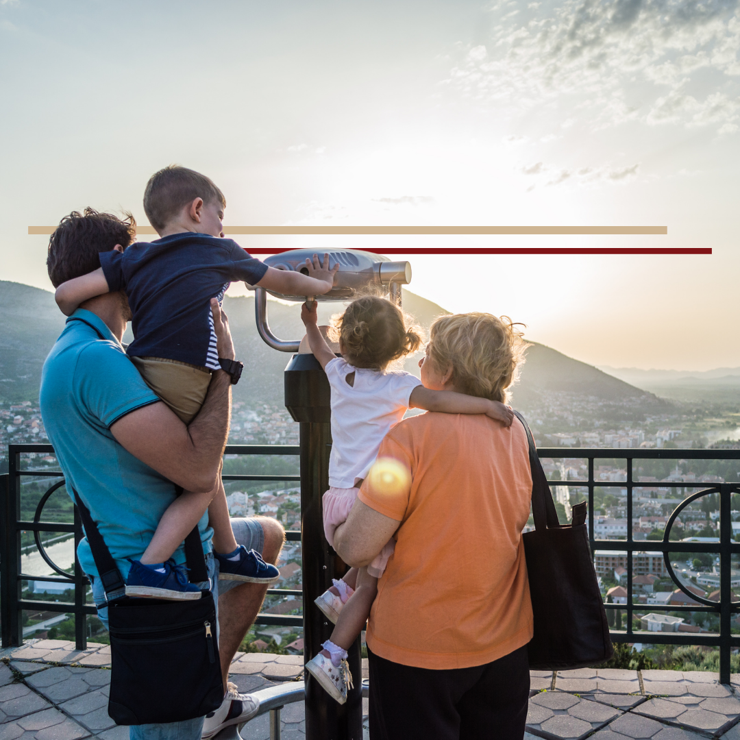 A family of 4 including a father and 2 preschool aged children with their grandmother looking through binoculars over a city in the distance at sunset